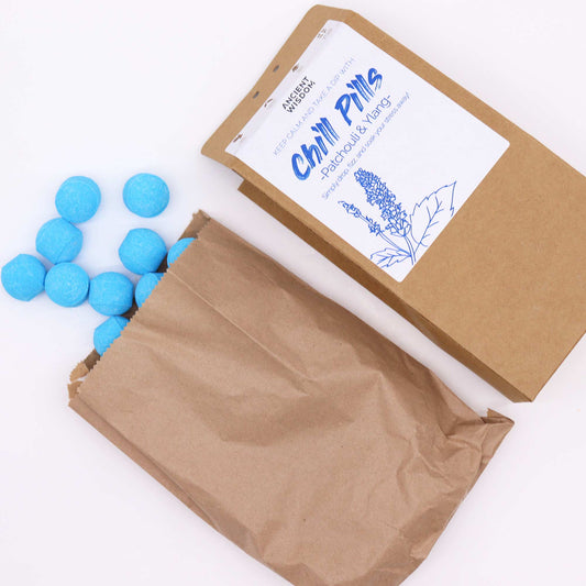 Brown paper bag labeled "Chill Pills," containing blue spherical tablets, spills onto a white surface. The label reads: "Ancient Wisdom. Keep Calm and Take a Dip with Chill Pills Patchouli & Ylang. Simply drop into a bath, sit back, and relax, and enjoy the amazing aroma!" A line drawing of a plant accompanies the text.