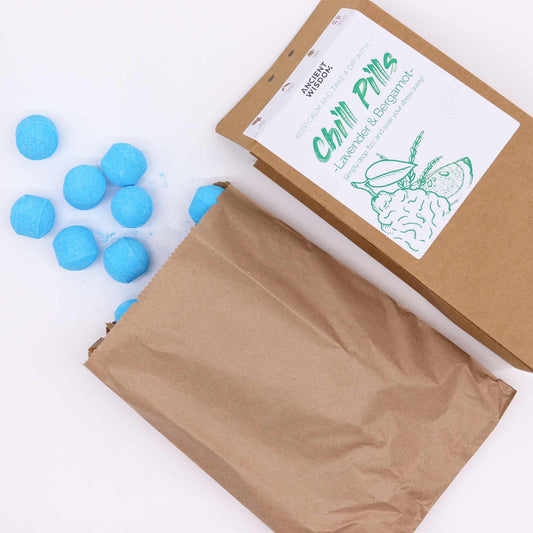 Blue spherical tablets spill from an open brown paper bag next to a package labeled "Chill Pills, Lavender & Bergamot, Ancient Wisdom." Set on a white surface.