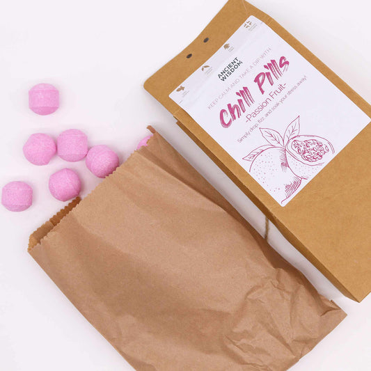 Pink, hexagonal candies spill from a brown paper bag beside a labeled package. Text: "ANCIENT WISDOM, Chill Pills, Passion Fruit. Simply drop, fizz, and soak your stress away."