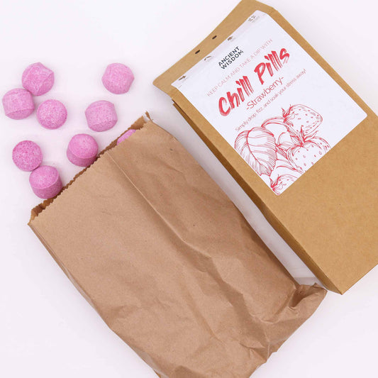 Brown paper bag spills pink, round bath fizzies beside labeled box reading: “Ancient Wisdom. Keep calm and take a dip with Chill Pills, Strawberry. Simply drop, fizz, and soak your stress away!”