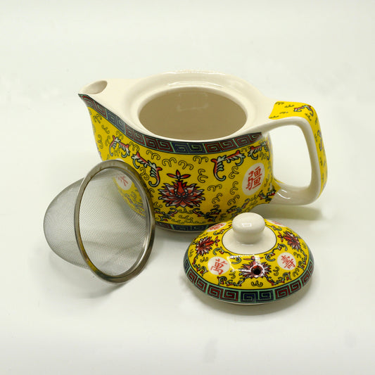 A yellow teapot with ornate detailing sits on a white surface, next to its lid and a metal tea strainer. The teapot's design includes red and blue floral patterns and Chinese characters.