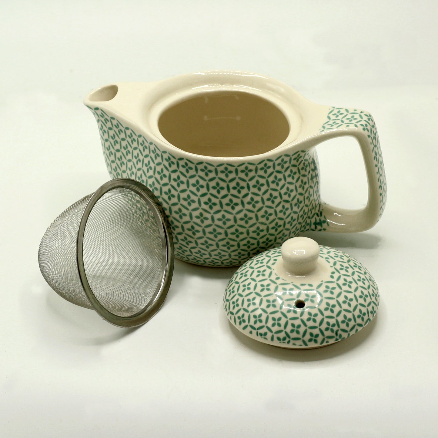 A ceramic teapot with a teal geometric pattern sits open, accompanied by its lid and a separate metal tea strainer, all on a white surface.