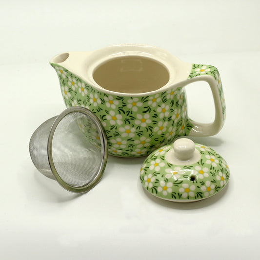 A green and white floral teapot sits with its lid and a metal infuser nearby on a white surface.