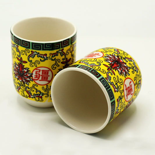 Two small, cylindrical ceramic cups, decorated with yellow and red floral patterns and Chinese characters, rest on a white surface.
