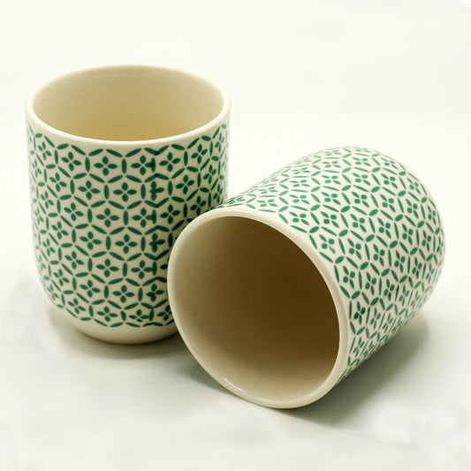 Two ceramic mugs, decorated with a green repeating pattern, rest on a white surface. One mug is upright; the other is lying on its side.