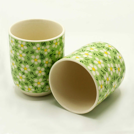 Two ceramic mugs, adorned with a light green daisy pattern, rest on a white surface. One mug is upright; the other is tilted, revealing its interior.
