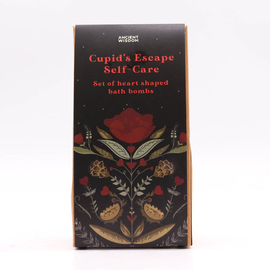A box with floral designs contains bath bombs. Text reads: "ANCIENT WISDOM Cupid's Escape Self-Care Set of heart shaped bath bombs." The design is on a black background with red flowers.