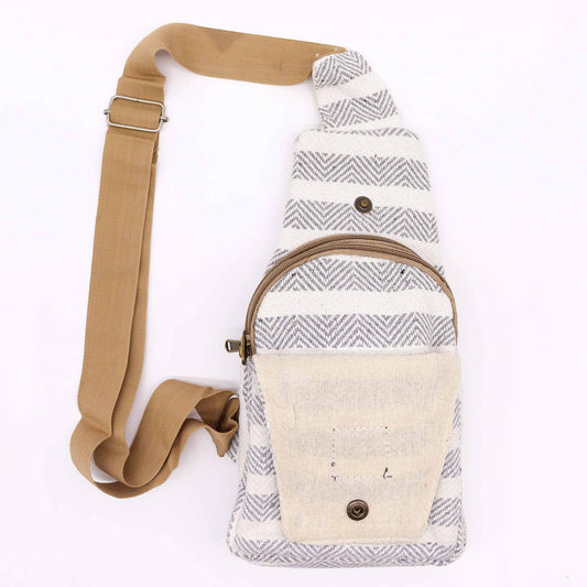 A patterned crossbody bag with a tan strap rests on a white surface. The bag has horizontal, gray, and white chevron stripes. It has a zippered pocket and a flap pocket with a snap closure.
