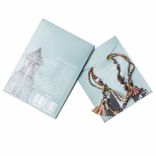 A bracelet with colorful tassels and beads sits atop a light blue card with text. Another identical card with a temple illustration and barcode text sits next to it: Wear the bracelet to find your belief and love of life.