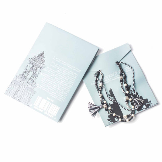 A beaded necklace lies on a pale blue card, beside a matching card featuring a sketched building and the text: "Wear this bracelet to understand the mystic balance good and evil..." plus barcode and "TSJ-03."