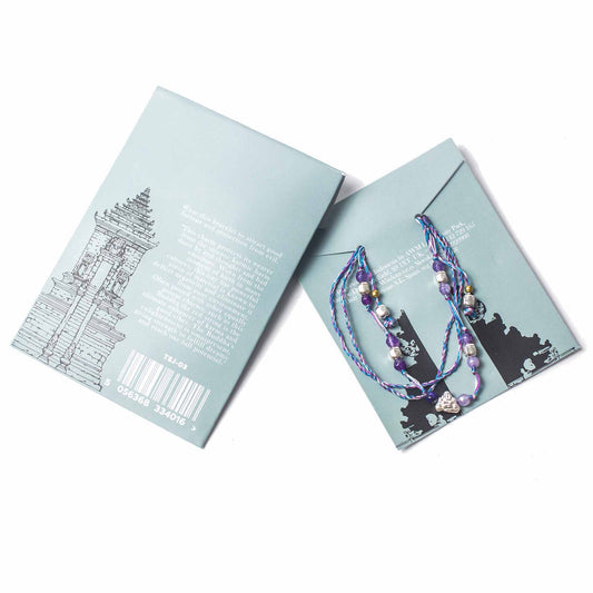 A bracelet is laid diagonally across two light blue product cards, one of which has a drawing of a temple and the text: "Wear this bracelet to attract good fortune and protection from evil."