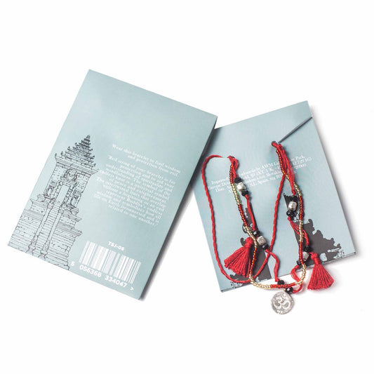 A beaded, red tassel bracelet with a metal "Om" charm sits on a blue product card, which has text including "Wear this bracelet to find wisdom and protection from evil" and a building sketch on the left side, on a white surface.