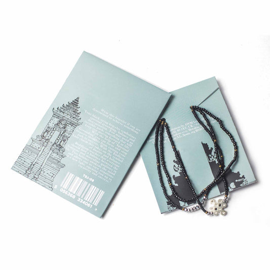 A black bead bracelet is placed atop two light blue cards on a white surface. The cards feature a temple illustration and text that reads: "Wear this bracelet if you are determined to achieve something...TSJ-08 5 056368 334061 >".