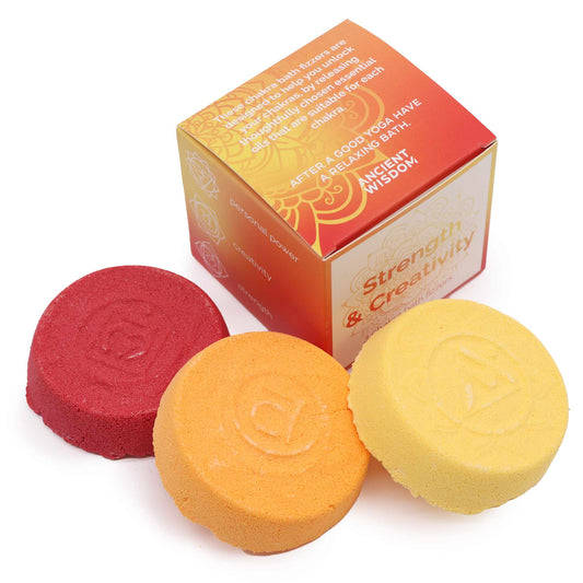 Three colored bath fizzers (red, orange, yellow) are placed next to a box labeled "Strength & Creativity." The box description reads: "These chakra bath fizzers are designed to help you unlock and activate your chakra energy by using thoughtfully chosen essential oils that are suitable for each chakra. After a good yoga have a relaxing bath. Ancient Wisdom."