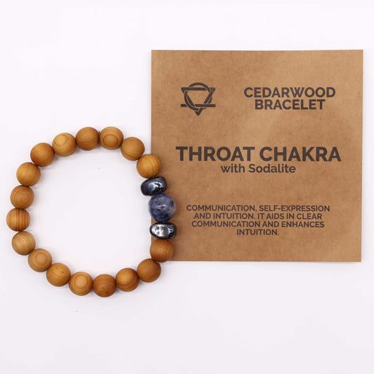 A beaded bracelet made of wood, silver, and sodalite lays near a small brown card on a white surface. The card reads: "CEDARWOOD BRACELET THROAT CHAKRA with Sodalite COMMUNICATION, SELF-EXPRESSION AND INTUITION. IT AIDS IN CLEAR COMMUNICATION AND ENHANCES INTUITION."