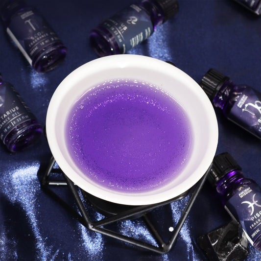 Purple liquid sits in a white bowl on a dark blue surface; surrounding it are several small bottles labeled with zodiac signs and the words "fragrance oil."