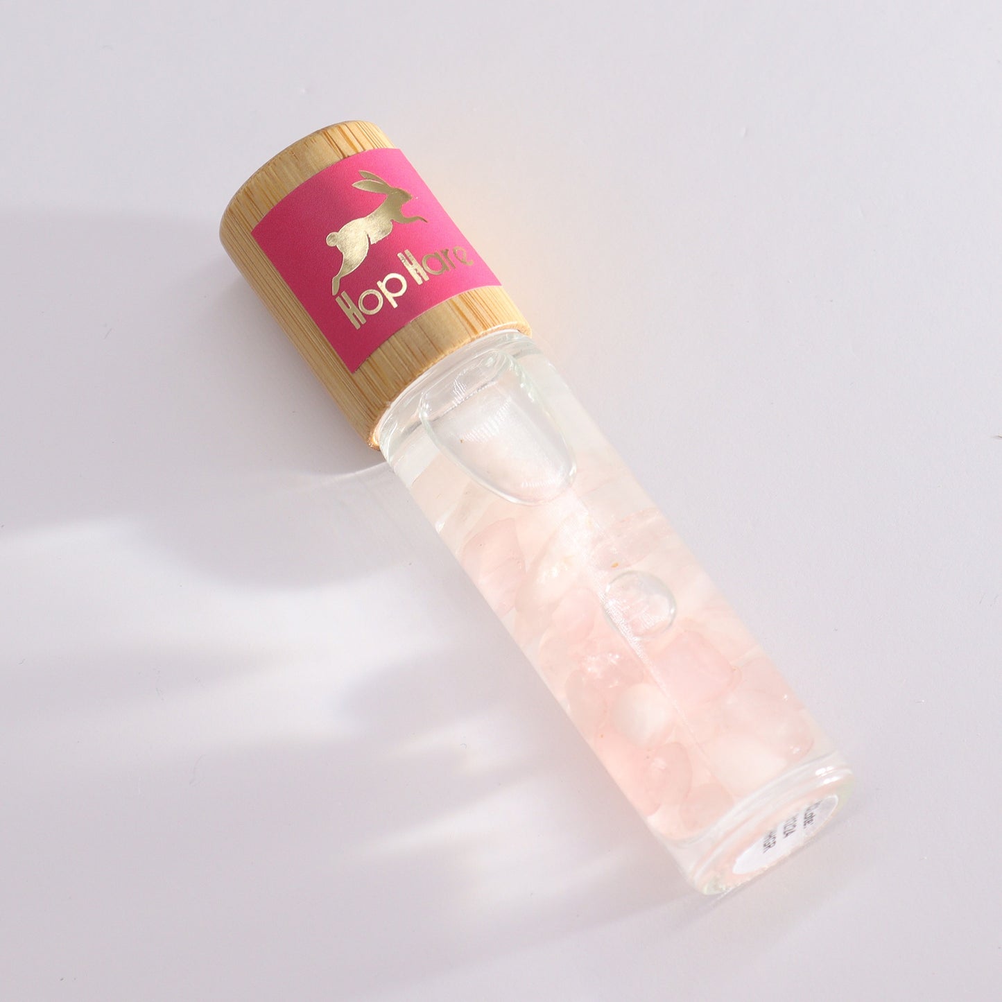 A glass roller bottle of serum, holding pink quartz, is on a white surface. The bottle has a bamboo top with a pink label that reads "Hop Hare" with an image of a rabbit.