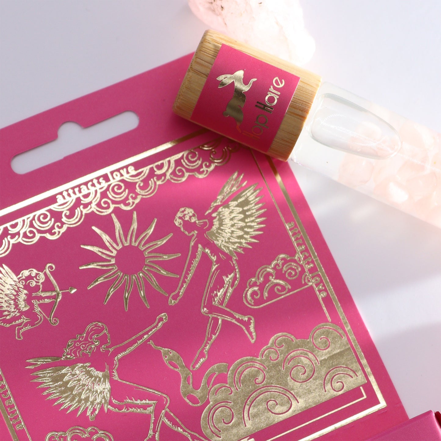 A roll-on fragrance bottle with a "lop hare" logo sits atop a pink card. The card features gold illustrations of angels and text that reads "attracts love."