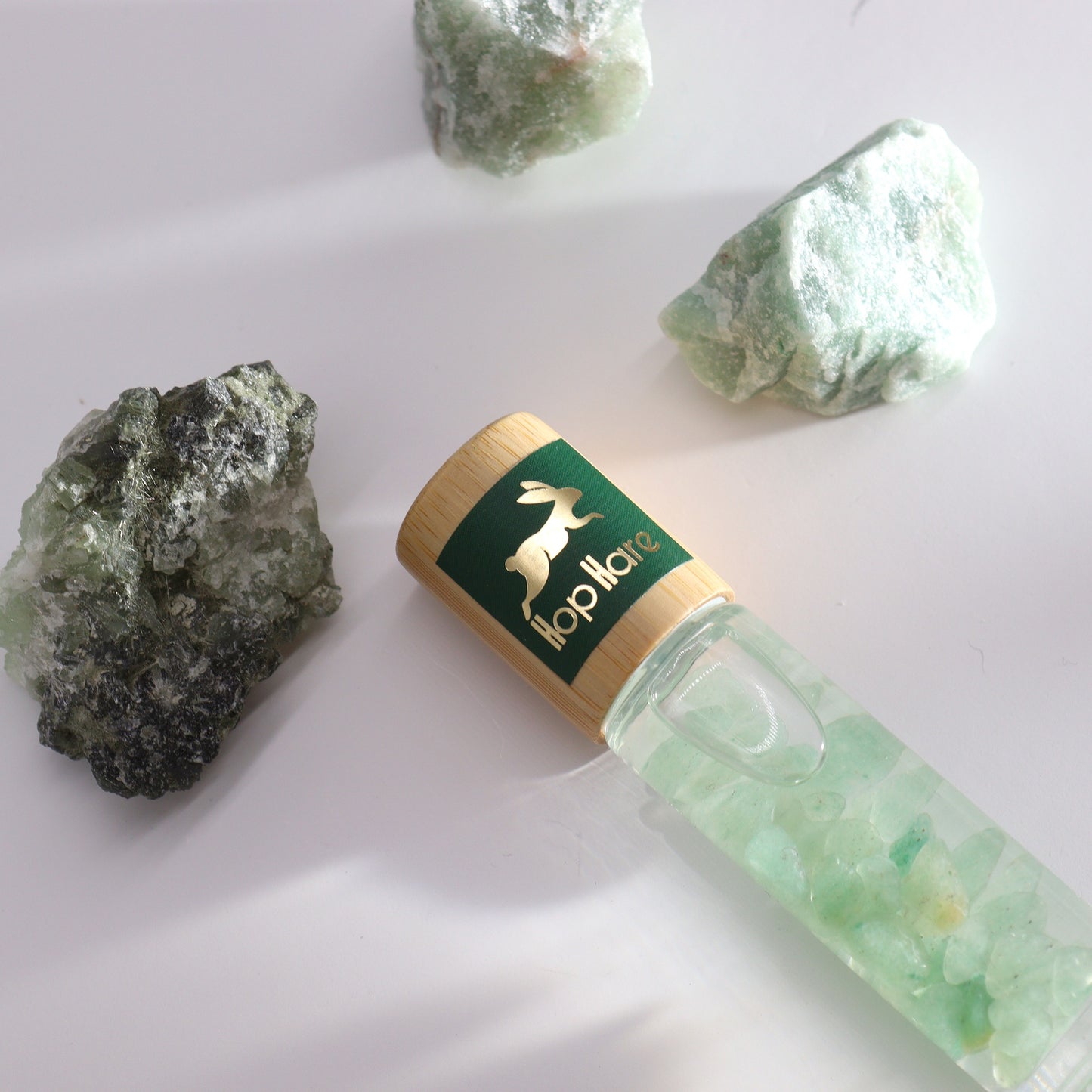 A clear bottle, filled with a clear liquid and small green crystals, lays on a white surface near three larger green rocks. The bottle's label reads "Hop Hare."