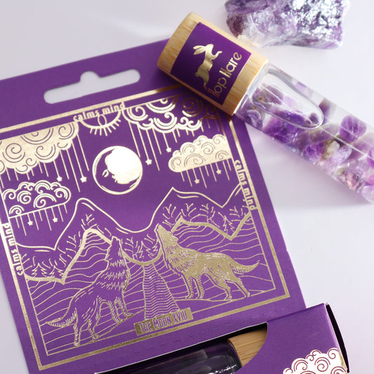 A purple box and oil roller, "lloplare," appear against a white background. The box illustration depicts two wolves howling at the moon over mountains. Text on the box includes: "calms mind," "THE MOON XVIII." The rollerball is filled with amethyst stones.