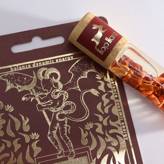 A small glass vial, labeled "hop Hare" and filled with orange bits, lays next to a burgundy card. The card features gold-foil art and the text "balance dynamic energy".