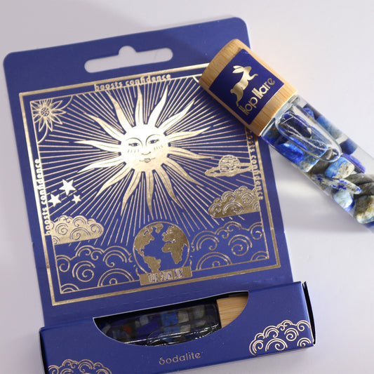 A lapis lazuli stone vial rests against its dark blue box. The box features gold foil art including a sun, planet, and clouds and reads "boosts confidence," "THE SUN XIX," and "Sodalite." The vial's wooden top reads "hop hare" with a rabbit silhouette.