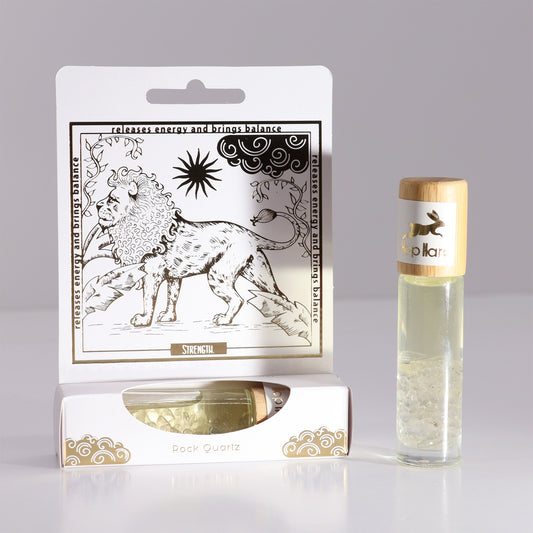 A clear glass rollerball bottle labeled "Hop Hara" and containing crystals stands next to its white branded packaging featuring a lion. Packaging text reads "releases energy and brings balance, STRENGTH, Rock Quartz".