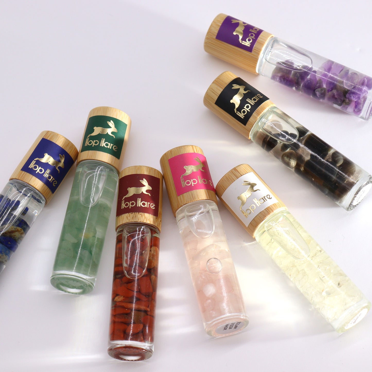 Several "Hop Hare" rollerball bottles, filled with oil and stones, are scattered on a white surface. The bottles have clear glass bodies and light wooden caps with colored labels; colors include green, pink, white, red, purple, blue and black.