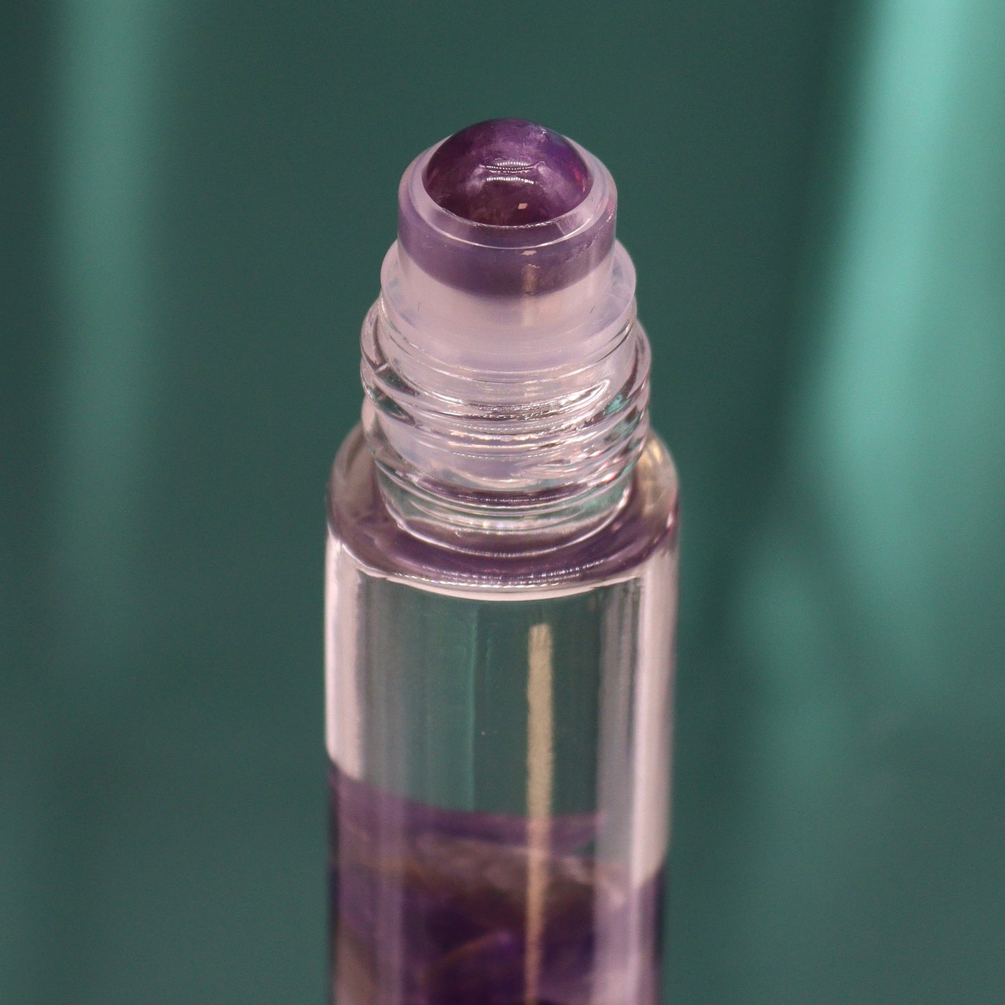 A clear rollerball bottle sits against a teal background. The bottle is filled with clear liquid and has purple crystal chunks at the bottom and a purple rollerball.