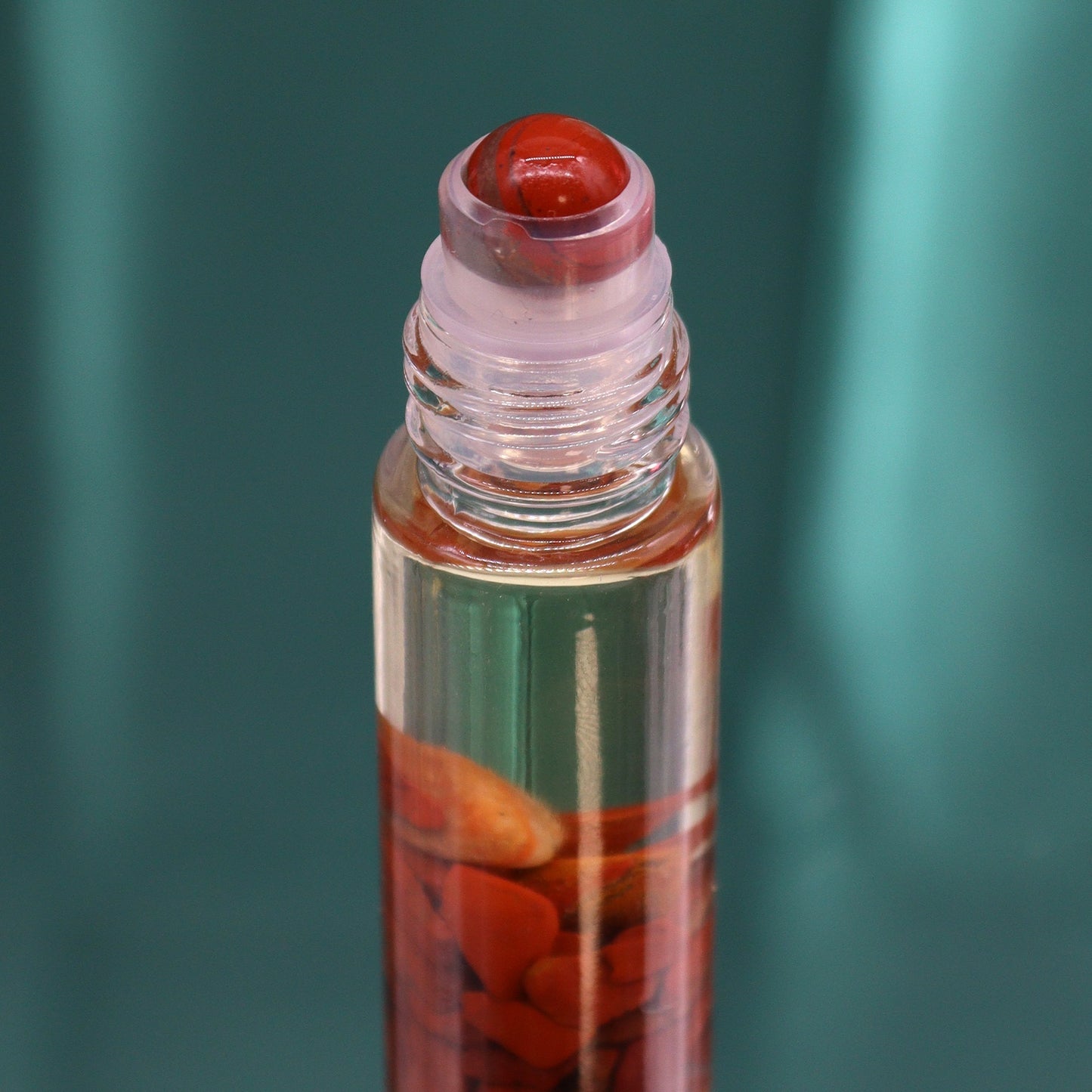 A clear glass roller bottle, partially filled with small red crystals, sits on a dark teal surface. The rollerball itself is also a red stone.