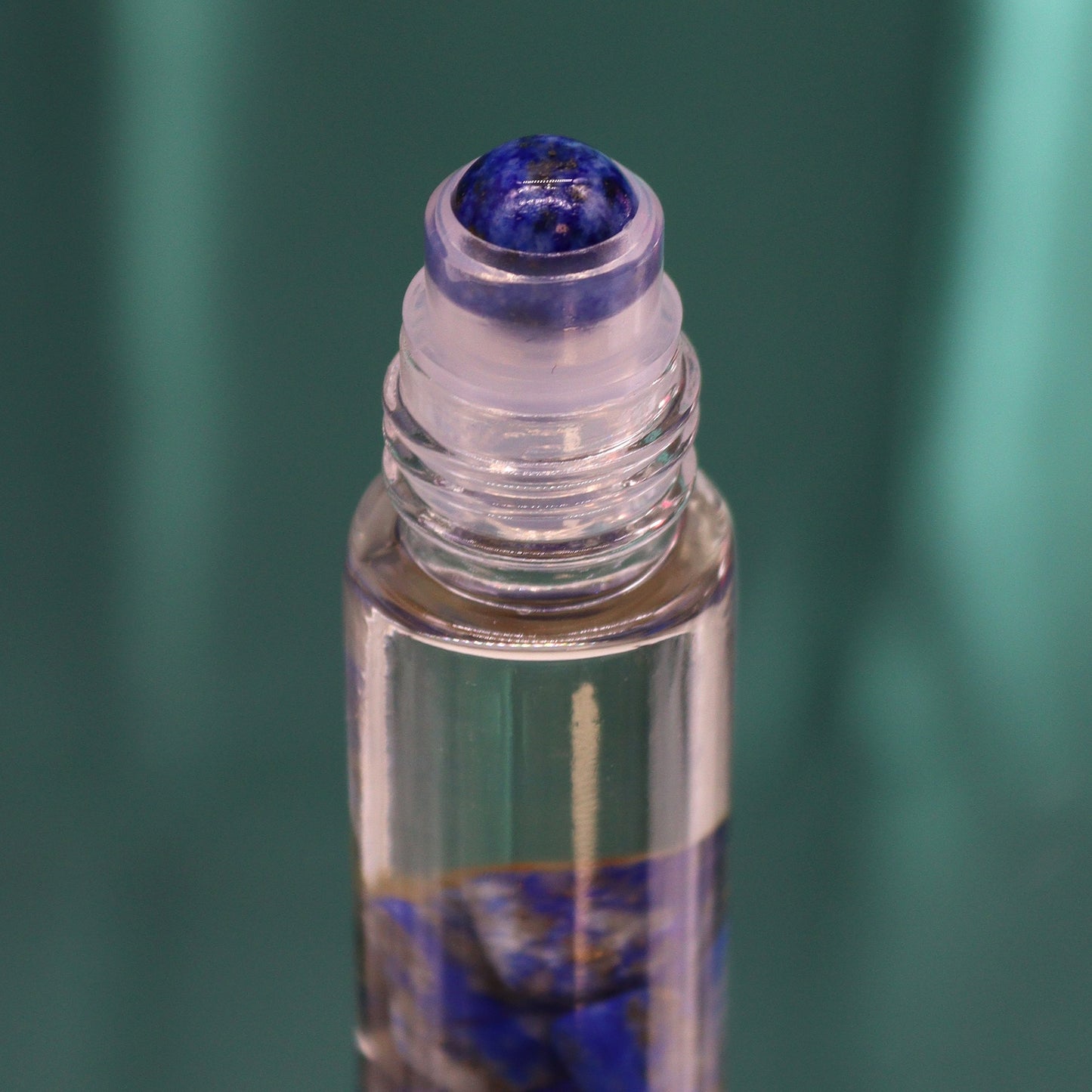 A clear glass vial containing blue stones and oil is displayed, with a blue rollerball top. The vial is centered against a blurred, teal background.