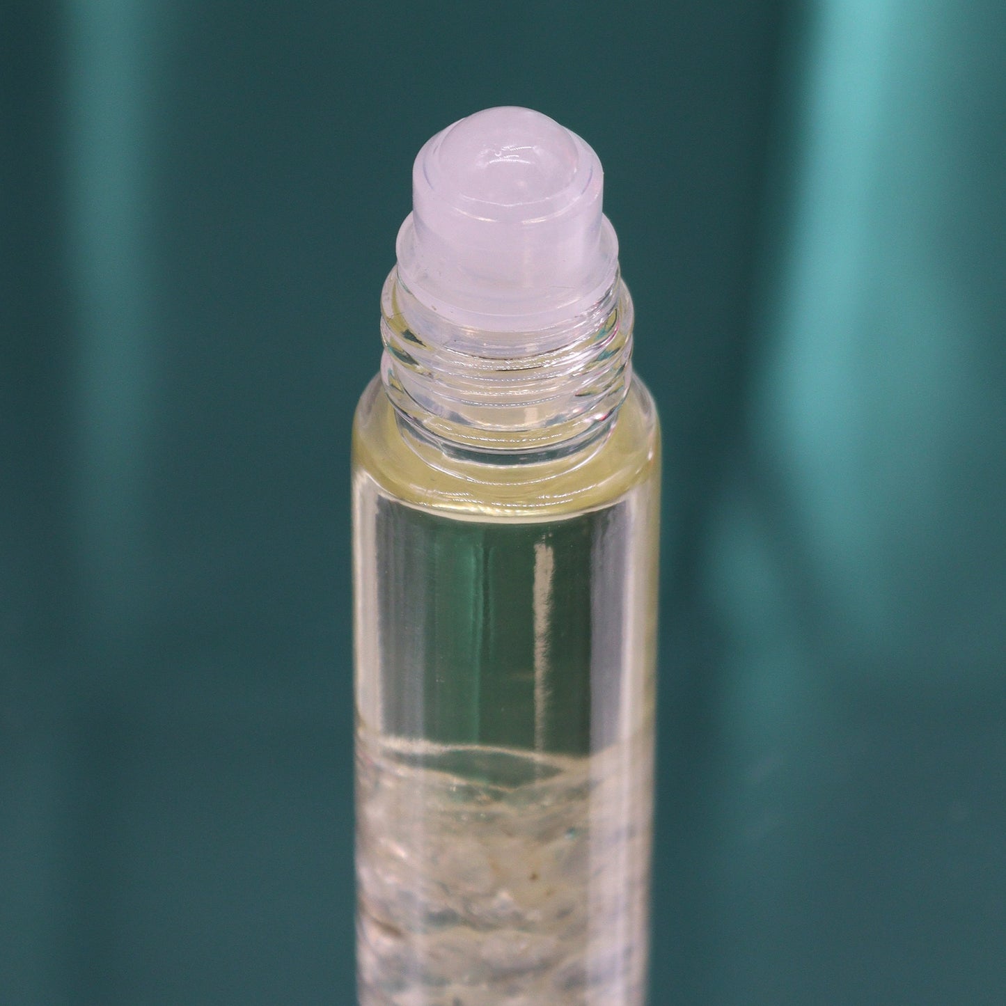 A transparent roll-on bottle stands on a teal surface, filled with a liquid. The top applicator is transparent.