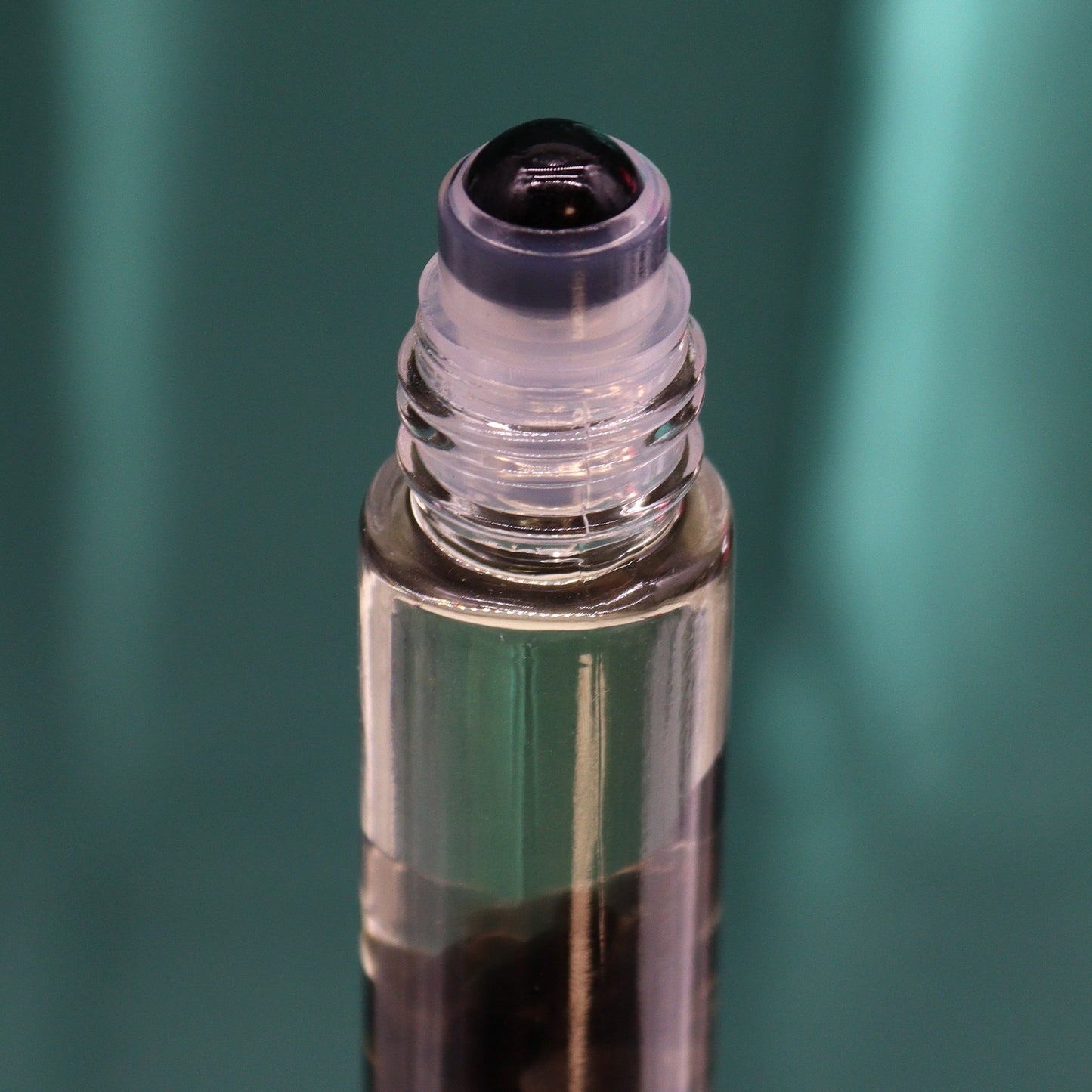 A glass roll-on bottle sits upright, filled with dark liquid. The bottle has a clear cap and a black rollerball and is placed against a teal background.