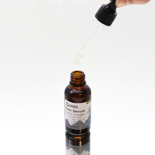 Brown glass bottle of hair serum receives a droplet from a held glass dropper over a white reflective surface; label reads: ANCIENT WISDOM Hair Serum Castor & Argan Fragrance Free For all hair types to nourish, strengthen and add shine to hair MADE IN ENGLAND 30ML 1.0FL OZ