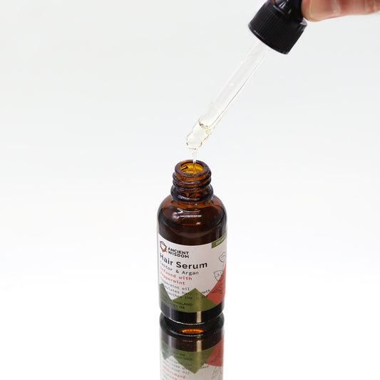 Amber glass hair serum bottle receives a droplet from a glass dropper above on a white reflective surface; label text: ANCIENT WISDOM Hair Serum Castor & Argan Infused with Peppermint Peppermint oil stimulates hair growth soothes the scalp Made in England 1 FL OZ ORGANIC