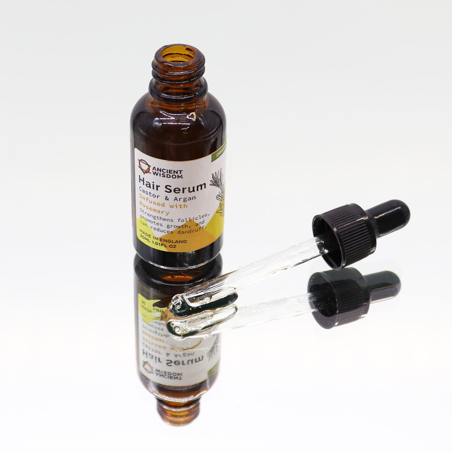 A brown glass bottle labeled "ANCIENT WISDOM. Hair Serum. Castor & Argan Infused with Rosemary. Strengthens follicles, promotes growth, and can reduces dandruff. MADE IN ENGLAND 30ML 1.01FL OZ" sits on a reflective surface with a glass dropper.