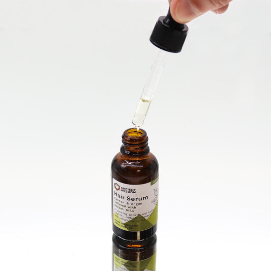 A brown glass bottle of Ancient Wisdom hair serum with a dropper releasing a clear oil drop into the open neck, resting on a reflective white surface; Label text: ANCIENT WISDOM Hair Serum Castor & Argan infused with Herbal Oils Promoting growth and scalp health, reducing dandruff and enhancing shine MADE IN ENGLAND 30ML 1.01FL OZ