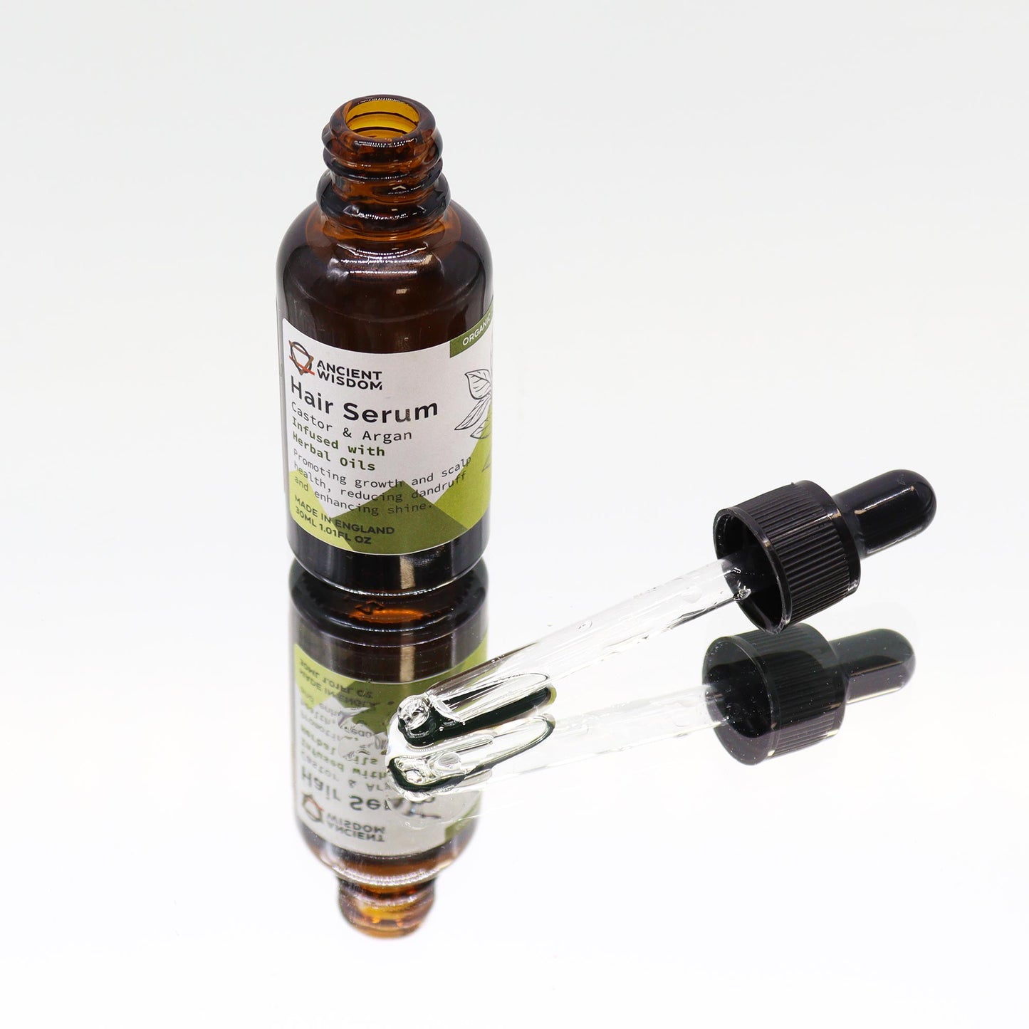 Amber glass bottle of hair serum sits open on a reflective surface beside a glass dropper; label reads: ANCIENT WISDOM Hair Serum Castor & Argan Infused with Herbal oils Promoting growth and scalp health, reducing dandruff and enhancing shine. MADE IN ENGLAND 30ML 1.01 FL OZ and a small green tag reading ORGANIC.