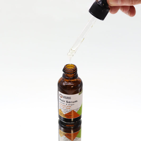 Brown glass hair serum bottle receives a drop of clear oil from a hand-held glass dropper above on a white reflective surface; label reads: ANCIENT WISDOM Hair Serum Castor & Argan Infused with Carrot Seed Rejuvenates and strengthens promoting growth enhancing shine MADE IN ENGLAND 30ML 1.01 FL.OZ ORGANIC