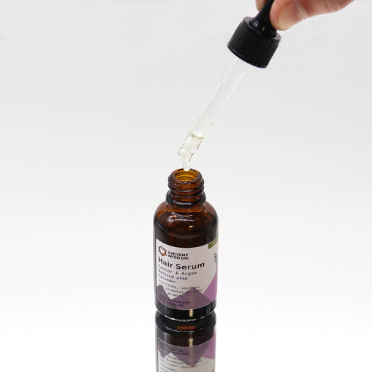 Amber glass hair serum bottle receives a clear drop from a held glass dropper above a white reflective surface; label text reads "ANCIENT WISDOM Hair Serum Castor & Argan Infused with Lavender nourishes, soothes, promotes healthy growth MADE IN ENGLAND 30ML 1.01FL OZ".