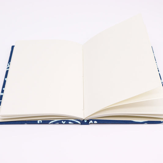 An open notebook rests flat; its blank, cream-colored pages are slightly turned. The cover, partially visible, features a blue pattern. The background is a plain white surface.