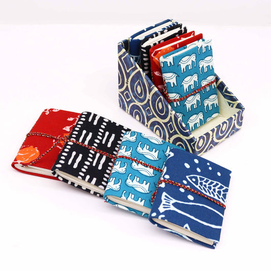 Colorful fabric-covered notebooks with animal and geometric prints are tied with cords. They are displayed in a matching, patterned holder, arranged on a white surface.