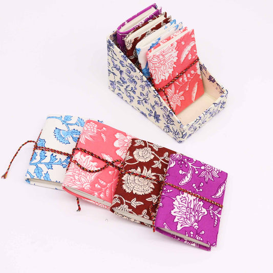 Colorful notebooks with floral patterns are displayed, some stacked vertically in a matching floral holder, while others lie horizontally, tied with cord, on a plain white background.
