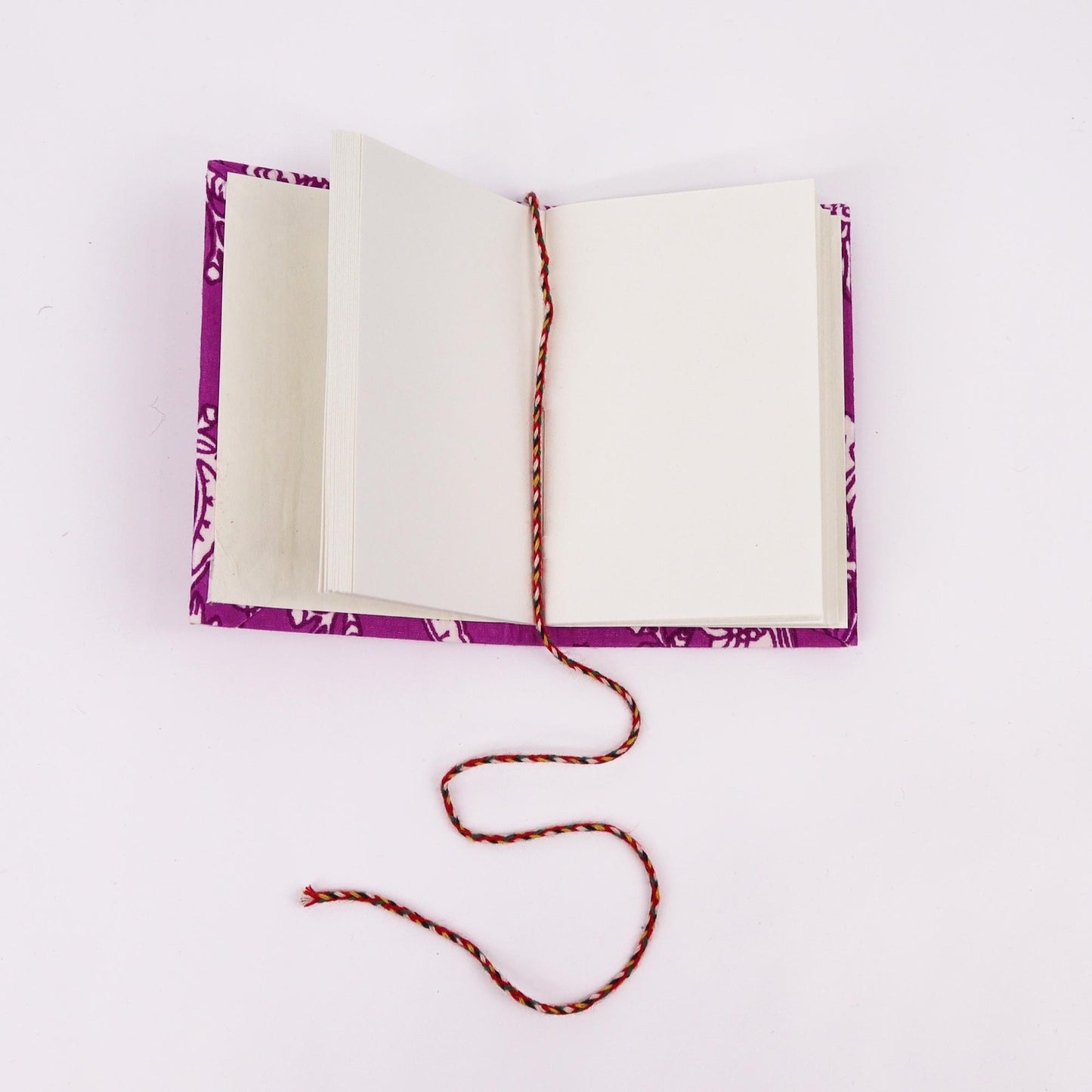 An open notebook with blank pages lies on a flat surface. A braided, multicolored string serves as a bookmark, partially draped across the pages. The cover features a purple floral pattern.