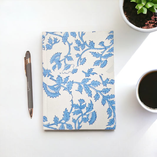 A notebook with a blue floral design lies on a white surface, accompanied by a gray pen, a potted plant, and a cup of coffee in the background.