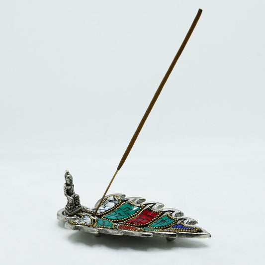A colorful, leaf-shaped incense holder supports a vertical incense stick, with a small, seated figure at one end. The setting is a plain, white background.