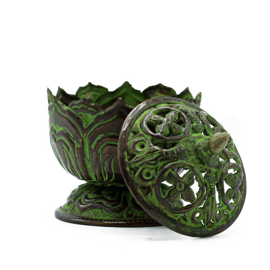 An ornamental green and bronze incense burner with intricate floral patterns is partially open. It rests on a pedestal, and the lid leans against it, revealing a pointed handle.