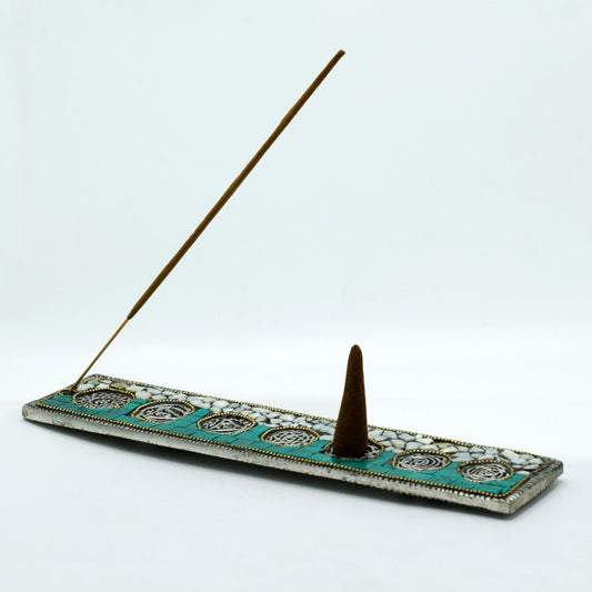 An incense holder features a burning stick and cone on a decorative, rectangular base with turquoise and white mosaic patterns, set against a plain white background.
