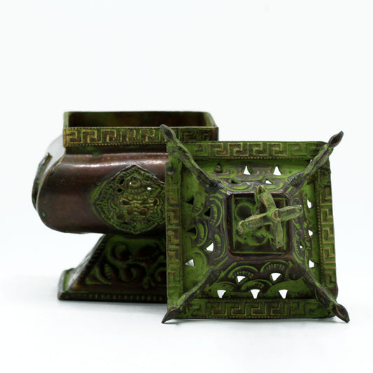 Intricately designed brass incense burner with a green patina, featuring geometric patterns and raised motifs. The lid is removed and placed beside it, showcasing detailed openwork.