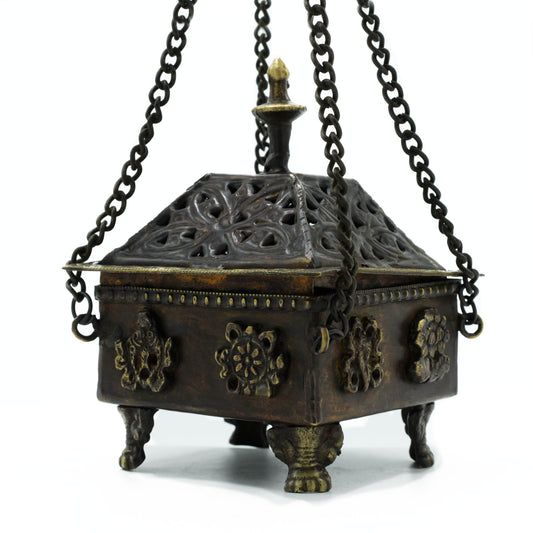 Ornate metal incense burner with intricate designs hangs from three chains. Supported by decorative feet, it features a pointed top and embossed floral patterns in a traditional setting.